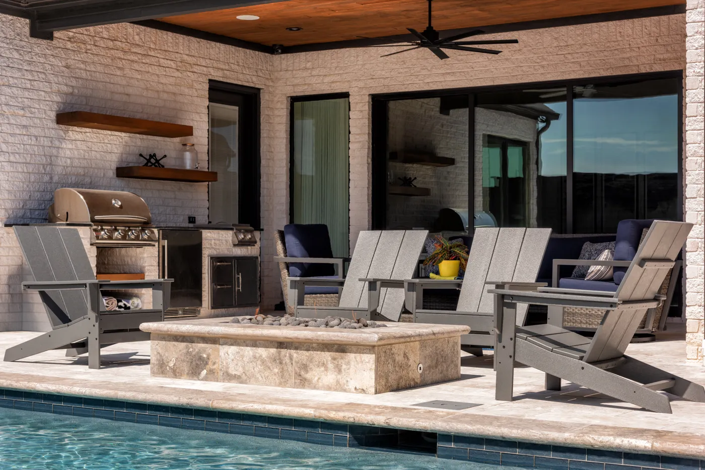 A covered outdoor kitchen and living area with a stone fire table, grill, and lounge chairs beside a pool.