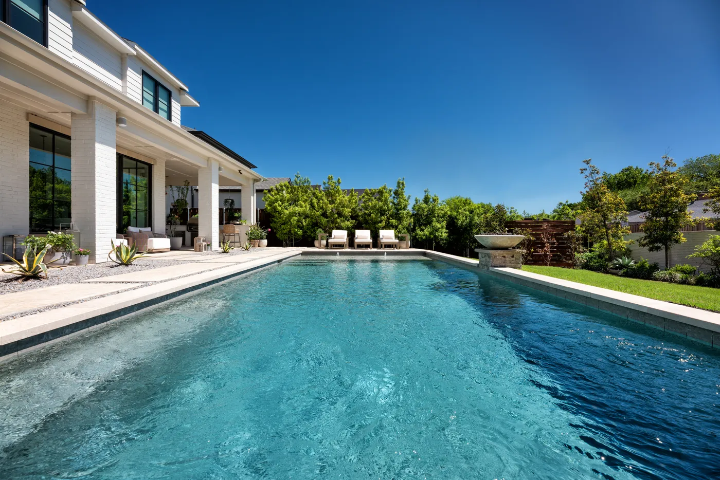 A long modern rectangular pool beside a white-brick two-story Texas home, with loungers and a stone water bowl.