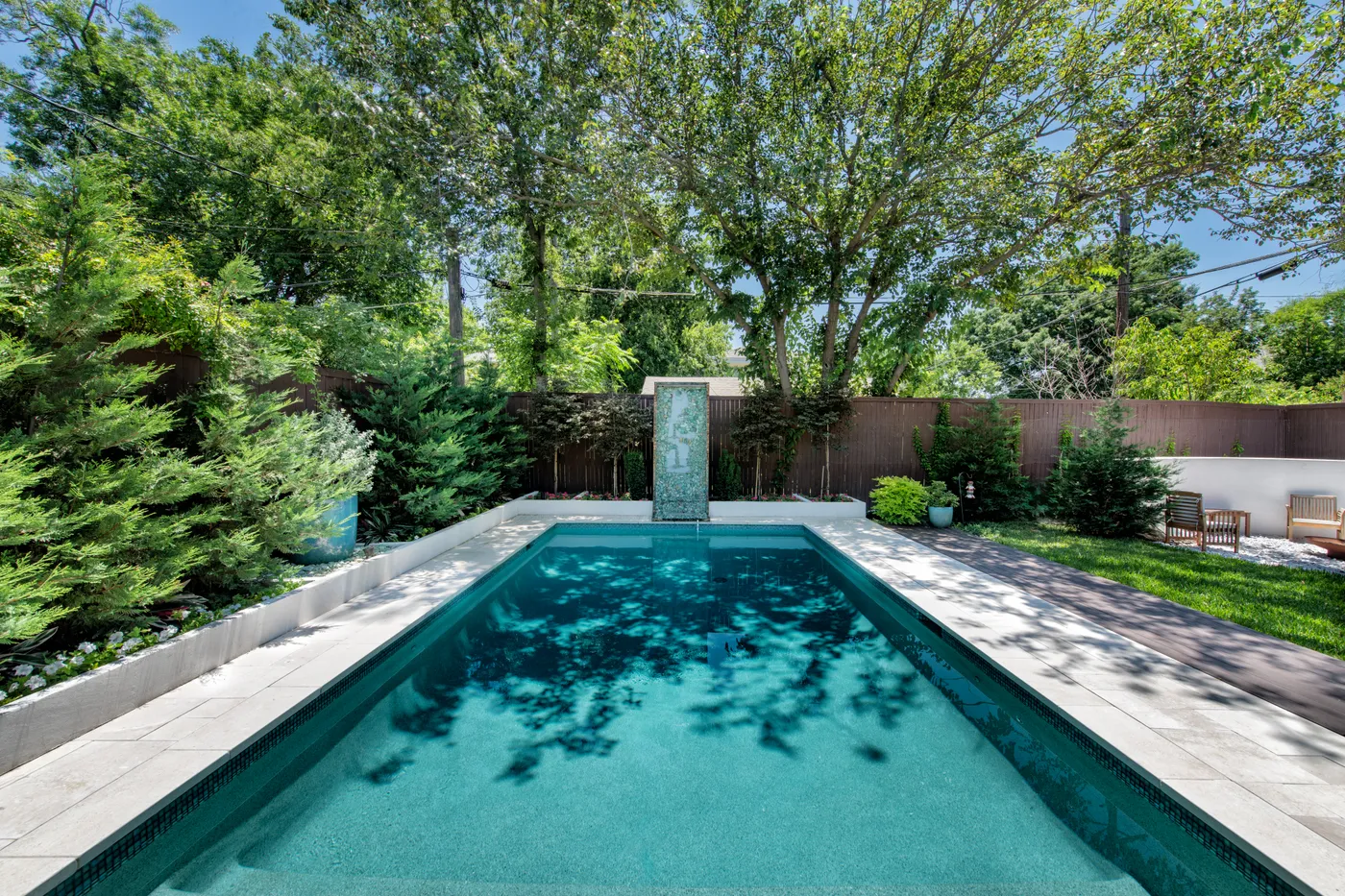 A rectangular pool with a glass water-feature column set in a lush, mature North Texas backyard.