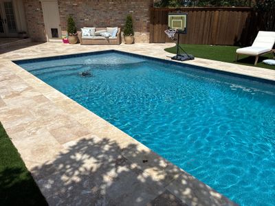 After: the same backyard rebuilt as a modern rectangular pool with travertine decking, turf, and a shaded lounge area.